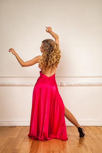 Rear view full length chic young woman in red dress posing at white wall background. Studio shot of cute blonde dancer woman with curly hair from behind. Dance psychology concept. Copy ad text space