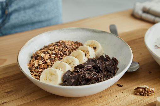 Healthy oatmeal for breakfast, with fresh organic banana, dark chocolate, nuts and grains, at a wooden table in a modern white kitchen, representing a healthy lifestyle, an image with a copy space