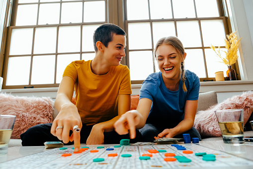 Two young women sitting on couch playing board game with counters, laughing. Happy couple bonding over an indoor leisure activity, smiling and sitting together at a table.