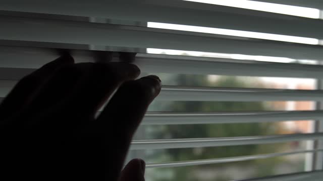 A curious man opens the roller blinds with his fingers and looks out the window.