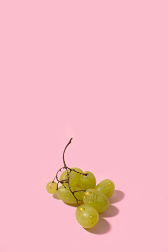 Branch of green grapes sprinkled with gold yellow shine glitter on a pink pastel background. Trendy minimal glamour concept. Autumn sparkle background.