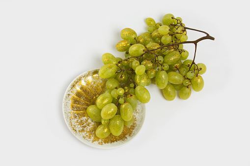 Branch of green grapes on a white plate sprinkled with gold yellow shine glitter on a white background. Trendy minimal glamour concept. Autumn sparkle background.