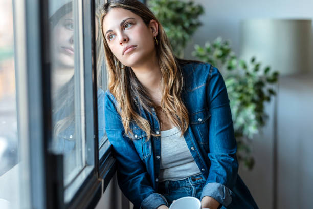 istock Beautiful young woman drinking a cup of coffee while looking forward standing next to the window at home. 1472765110