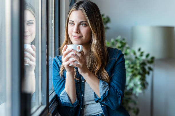 istock Beautiful young woman drinking a cup of coffee while looking forward standing next to the window at home. 1472765112