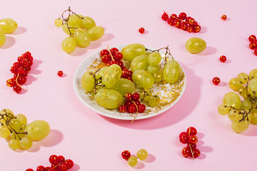 Branch of red currants and white grapes on a white plate sprinkled with gold glitter on a pink background. Trendy minimal creative concept.