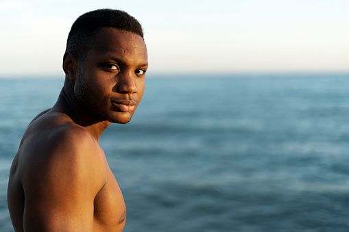 Handsome young multiracial man with naked torso standing on the beach and looking at the camera while warming at the sun