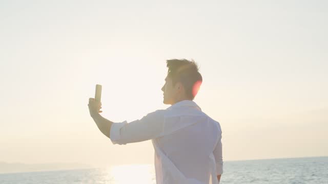 Asian young man tourist using smartphone taking photo during yachting. Attactive male feel happy and relax while sitting alone on deck of yacht, enjoy time when catamaran boat sailing during summer.