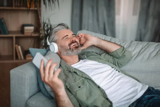 Photo of Glad caucasian old man with beard in headphones lies on sofa enjoys favorite music in smartphone in living room