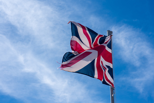 The flag of the United Kingdom of Great Britain and Northern Ireland, known as Union Flag or Union Jack, hanging down loosely at full-mast on a white pole against blue sky.