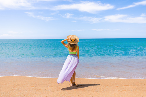 Attractive woman in straw hat and waving skirt standing on sandy beach, enjoy turquoise ocean and sunny skies on tropical vacation, exotic location. Back view.