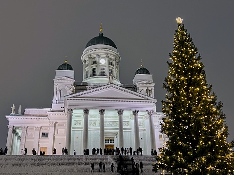 Winter twilight view to the front side of the Helsinki Tuomionkirkko white lighted cathedral with public on the stairs and Christmas tree with plenty lights and shining star on the top