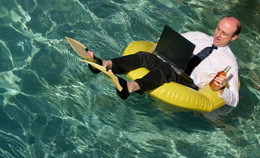 Businessman in suit and tie and working on laptop with a beer in one hand wearing flippers and floating on an inflatable boat in the crystal clear ocean