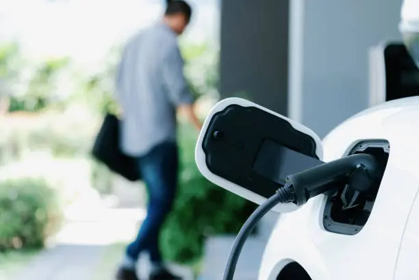 Focus electric car charging at home charging station with blurred progressive man walking in the background. Electric car using renewable clean for eco-friendly concept.