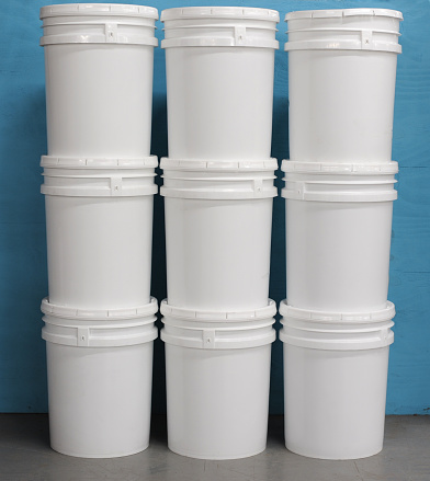 Nine white, plastic and unmarked buckets with lids are stacked in three columns against a blue dividing wall.