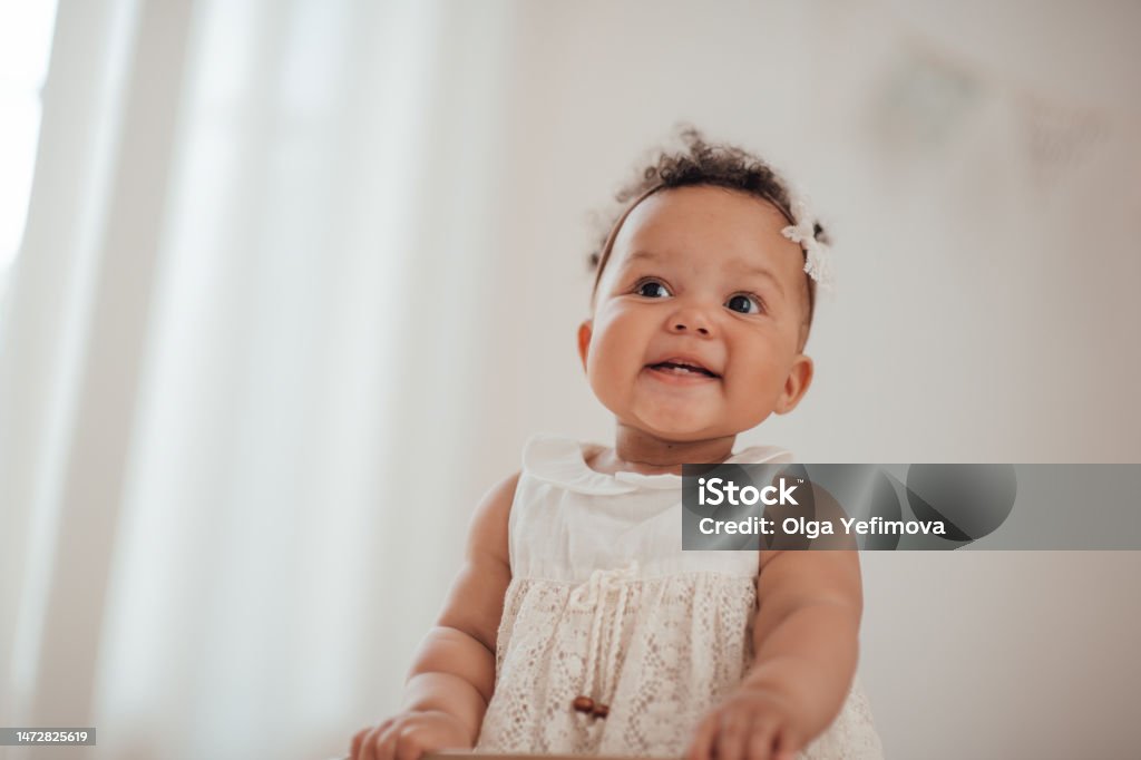 Portrait of mixed race toddler girl in round wooden baby crib at cozy childrens room neutral tones Candid authentic portrait of cute african american diverse female child at home interior of earth colors. Happy swarthy infant baby stands in kids bed in white lace dress and bow headband at bedroom Baby - Human Age Stock Photo
