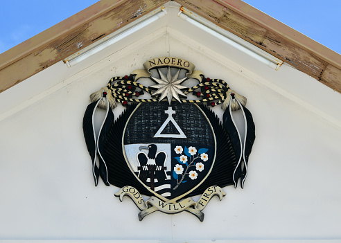 Yaren, Nauru: Coat of arms of the Republic of Nauru, tympanum of the Parliament building. The shield is divided and separated in the middle. In the upper section the alchemical symbol of phosphorus is shown over a woven background. The lower silver section depicts a black frigatebird, which sits on a perch over blue ocean waves. The lower right section is blue and contains a branch of calophyllum flowers. The shield is surrounded by images of tribal chief gear - ropes from palm leaves, feathers of the frigatebird and shark teeth. The twelve-pointed star above the shield is taken from the flag. The ribbon above it bears the name of the island in Micronesian Nauruan: \