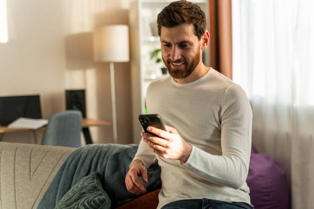 istock Handsome young male smiling while sitting with smartphone and looking at the screen with happy face 1472829466