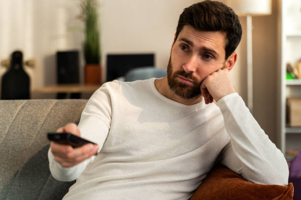 istock Bored young caucasian man holding tv remote control while sitting on a couch at home 1472829519