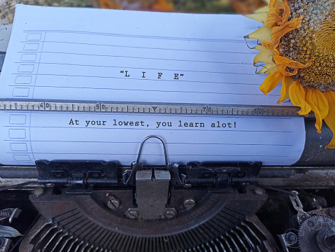 Life inspirational quote - At your lowest, you learn a lot! With typewriting text on white line paper on an old retro typewriter and wilted sunflower vintage. Life story concept - learn from mistakes.