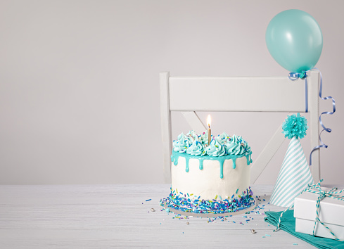 Blue and white birthday party with a buttercream drip cake, sprinkles, bright gold candle, hat and balloons on a light grey background. Trendy. Copy space.