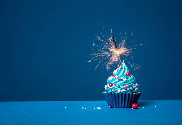 Blue cupcake with red and white sprinkles and lit sparkler on a blue background. Festive buttercream birthday cupcake with red, white and blue sprinkles and lit sparkler on a dark blue background. cupcake candle stock pictures, royalty-free photos & images