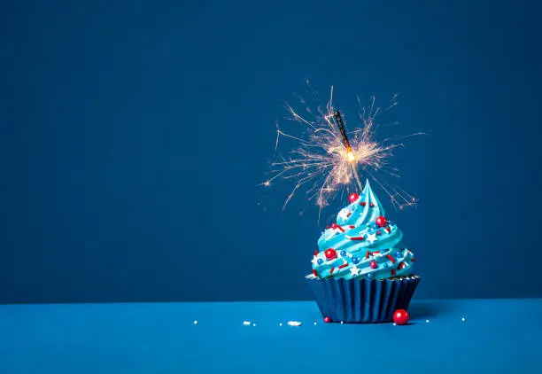 Festive buttercream birthday cupcake with red, white and blue sprinkles and lit sparkler on a dark blue background.