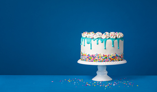White Birthday drip cake with teal ganache and colorful sprinkles over a dark blue background. Simple and trendy. Copy space.