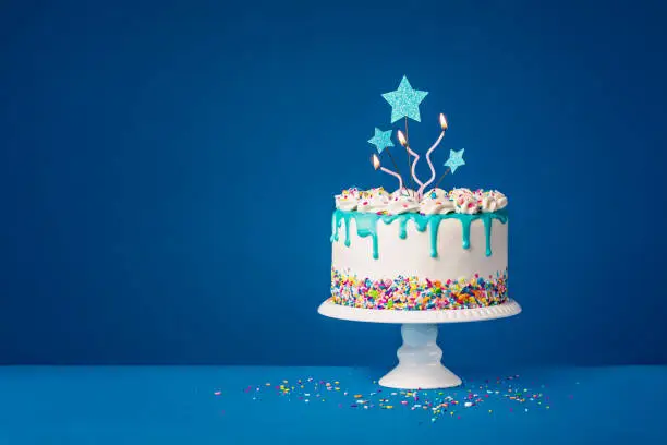 White Birthday cake with trendy teal ganache drips and colorful sprinkles, star toppers and fun candles over a dark blue background. Copy space.