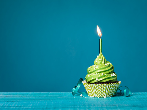 Buttercream birthday cupcake with green icing, lit candle, sprinkles, and ribbon on a teal blue background.