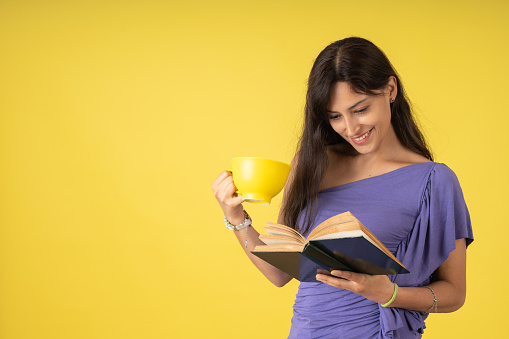 Young woman in a purple polo shirt happily reading an antique book while drinking a cup of tea, on a yellow background