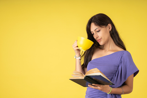 Very interested young woman reading a book while drinking a cup of coffee, on a yellow background