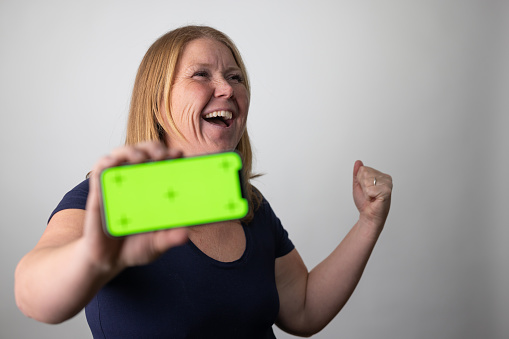 A woman celebrating against a grey studio background holding a smart phone