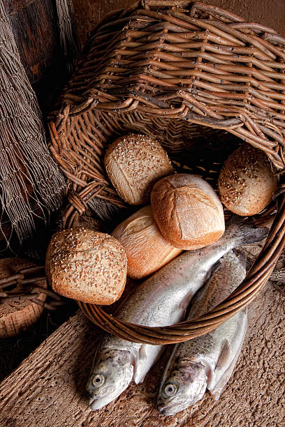 An overturned basket of bread loaves and two uncooked fish Religious still life of loaves of bread, fishing net, basket and two fresh fish loaf of bread stock pictures, royalty-free photos & images