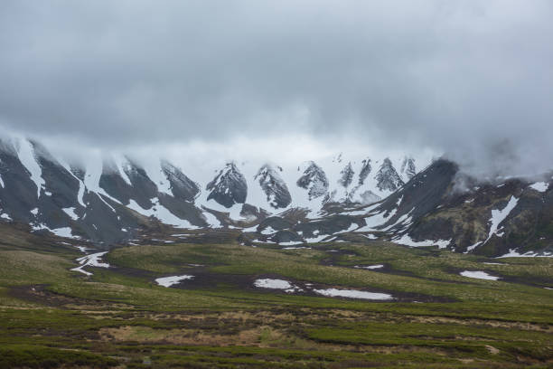 Dramatic alpine landscape with snowy mountains in gray low clouds. Bleak atmospheric scenery of tundra under lead gray sky. Gloomy minimalist view to mountain range among low rainy clouds in overcast.