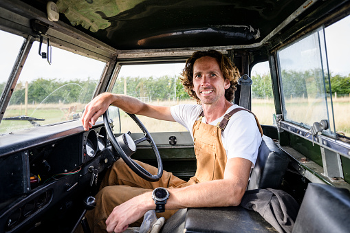 View through passenger window of Caucasian man in bib overalls sitting in driver’s seat of old farm vehicle and smiling at camera.