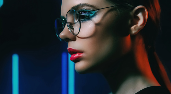 Retro look. Eyewear fashion. 2000s beauty. Blue color neon light closeup profile portrait of confident teen girl model face in glasses with artistic makeup on dark.