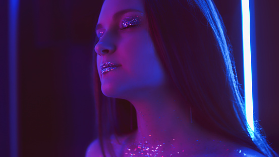 Neon girl. Glitter makeup. Nightclub fashion. Purple blue color glow relaxed woman with sparkling face skin eyeshadow lips in blur fluorescent light on dark free space.