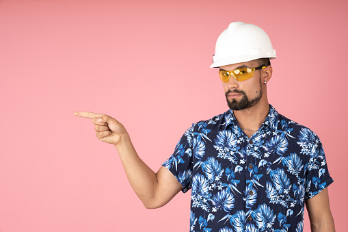 Man with helmet and goggles pointing to the left, on a pink background