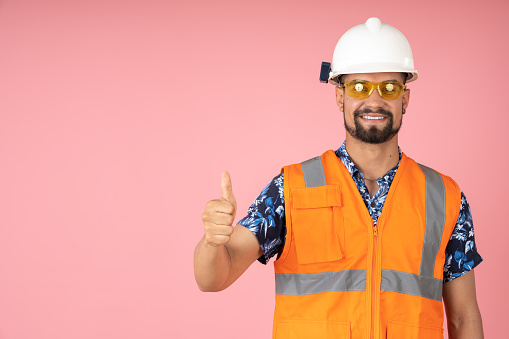 Worker with reflective vest, pointing his thumb up in agreement, on a pink background.