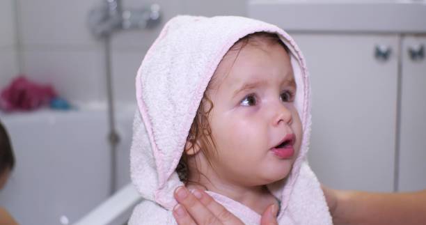 A beautiful baby girl stands in a towel in the bathroom and looks at her mother cutely. The child is dried with a towel after bathing. The concept of caring and caring for a child. A beautiful baby girl stands in a towel in the bathroom and looks at her mother cutely. The child is dried with a towel after bathing. The concept of caring and caring for a child.. cutely stock pictures, royalty-free photos & images