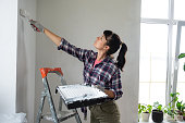 Woman in paint roller and white paint paints the wall of house on stepladder. Construction work and cosmetic repairs in house, wall painting, tinting, finishing work with your own hands