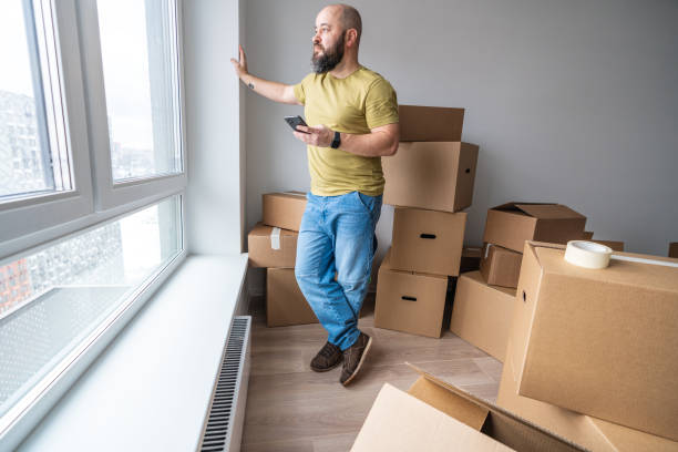 A man stands at the window in the room full of cardboard boxes and looking to a view, holding smartphone in the hand