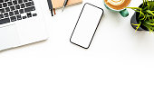 Smartphone with blank empty mockup screen is on top of white office desk table with laptop computer and supplies. Top view with copy space, flat lay.