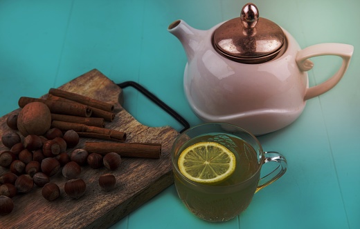 side view of lemon juice with lemon slice in glass cup and nuts cinnamon walnut on cutting board with teapot on blue background
