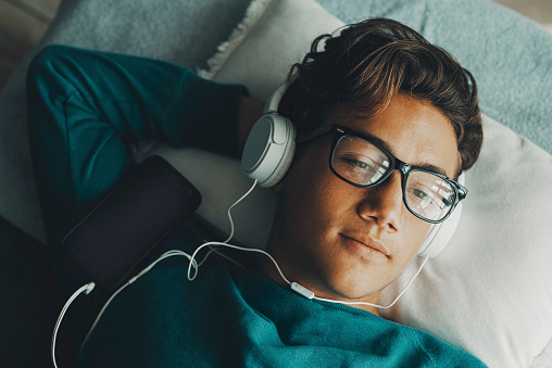 Handsome teenager above view asleep and relaxing with playlist music and headphones. Modern young guy using cellphone to listen podcast or school class alone at home laying on sofa. Relax afternoon