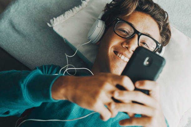 Cheerful young boy using phone app to listen music with headphones. Modern people chatting and dating online with cellphone device. Above view of student enjoying surfing net internet activity leisure stock photo