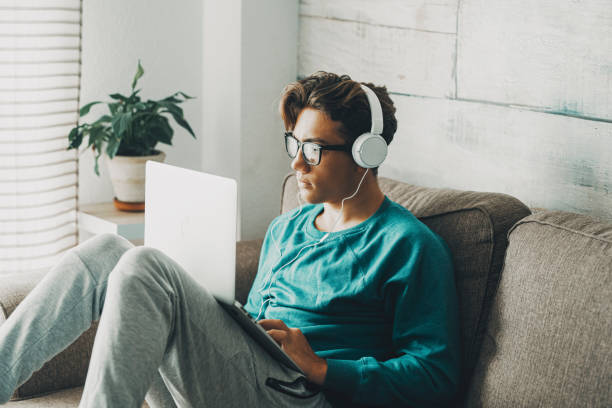 Relaxed young teenager using computer sitting on the sofa at home for play or study. Wearing headphones. One teenager boy listening music in indoor relax leisure activity alone. Afternoon time alone stock photo