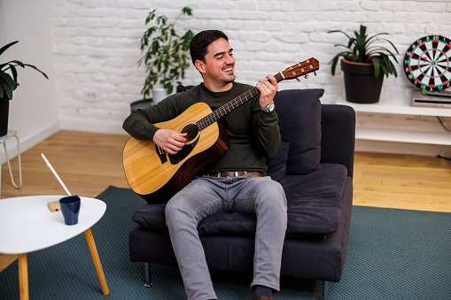 A young man is sitting on a sofa in the living room and playing an acoustic guitar and enjoying the sounds of the music.