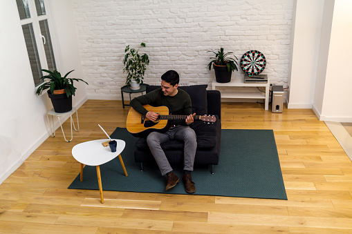 A young man is sitting on a sofa in the living room and playing an acoustic guitar and enjoying the sounds of the music.