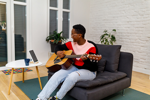 A young African woman is playing an acoustic guitar in the living room of the modern apartment.